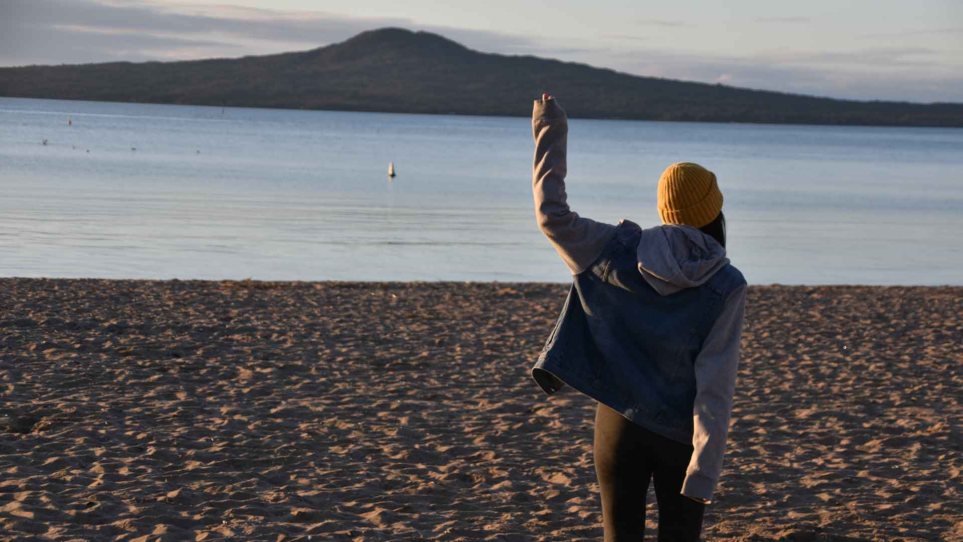 Camila abrigada en la playa con la silueta de Rangitoto al fondo